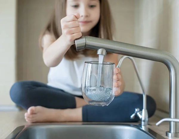 Little girl open a water tap with her hand holding a transparent glass. Kitchen faucet. Filling cup beverage. Pouring fresh drink. Hydration. Healthcare. Healthy lifestyle. World water day