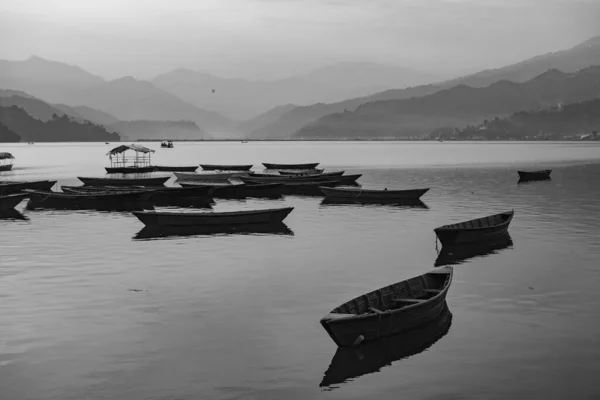 View over a few wooden boats on Phewa Lake in Pokhara, Nepal with the surrounding mountains in the background in black and white. Clear sunny day. Relaxation and calm serenity, peaceful and quiet.