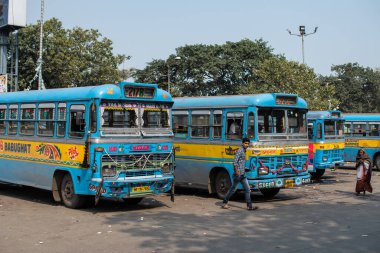 Kolkata, Hindistan - 1 Şubat 2020: Kimliği belirsiz bir erkek ve kadın 1 Şubat 2020 tarihinde Kolkata, Hindistan 'da bir otobüs durağında park etmiş birkaç geleneksel turkuaz ve sarı halk otobüsünün yanından geçtiler.
