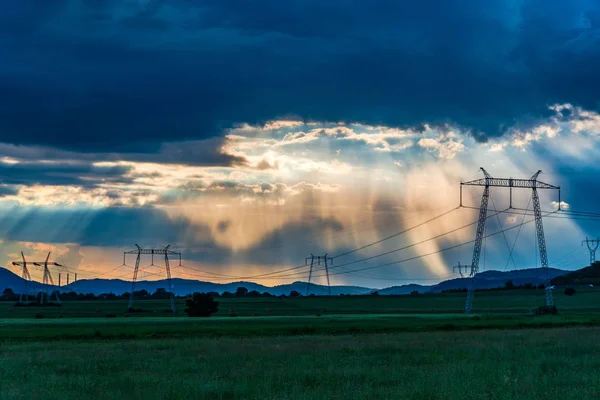 cloudy sunset sky with sunbeams and electric poles towers in field