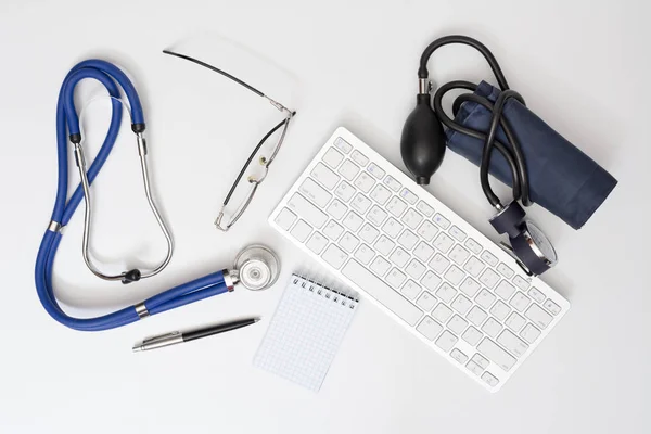 Stethoskop und Tonometer auf dem Tisch in der Arztpraxis. Draufsicht auf den Schreibtisch mit drahtloser Computertastatur, Brille und Notizbuch mit Stift — Stockfoto