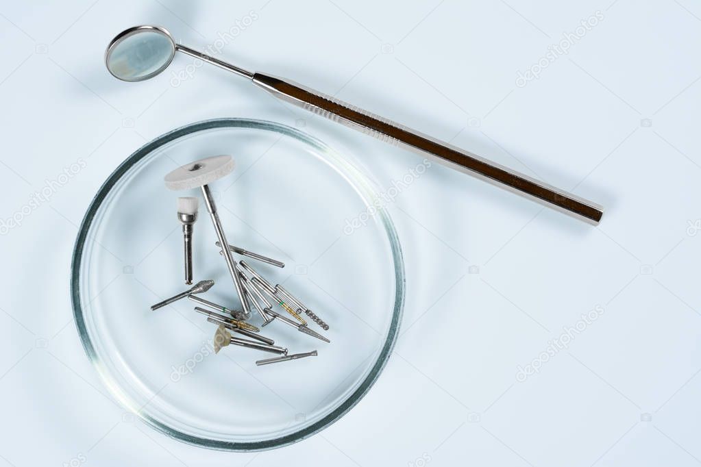 Dental medical tools, dentist equipment: stomatological mirror and used dental burs in Petri dish glass on light blue dentist table.