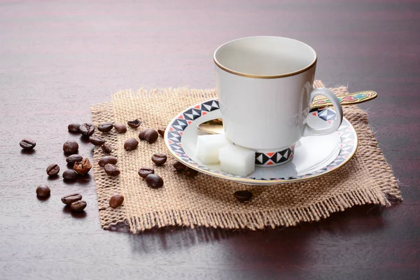 Porcelain coffee cup with saucer and golden spoon on a jute napkin. Vintage coffee set on wooden table with sugar and dropped coffee beans — Stock Photo, Image