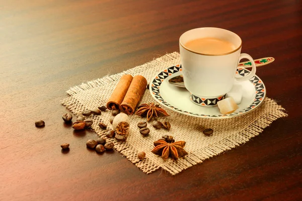 Coffee in a porcelain cup with a saucer, cinnamon, nutmeg and star anise on a jute napkin. Vintage coffee set on wooden table with scattered coffee beans. — Stock Photo, Image