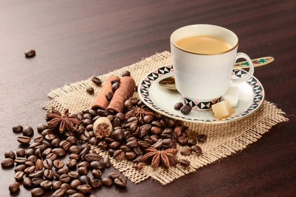 Coffee in porcelain cup on saucer with cinnamon, nutmeg and star anise on jute napkin. Vintage coffee set on wood table with scattered coffee beans. — Stock Photo, Image