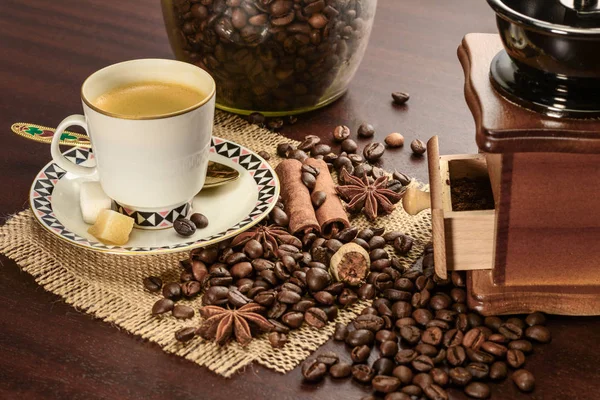 Cup of coffeee with saucer on a jute napkin. Vintage still life on wooden table with sugar, anise, cinnamon, grinder and coffee jar — Stock Photo, Image