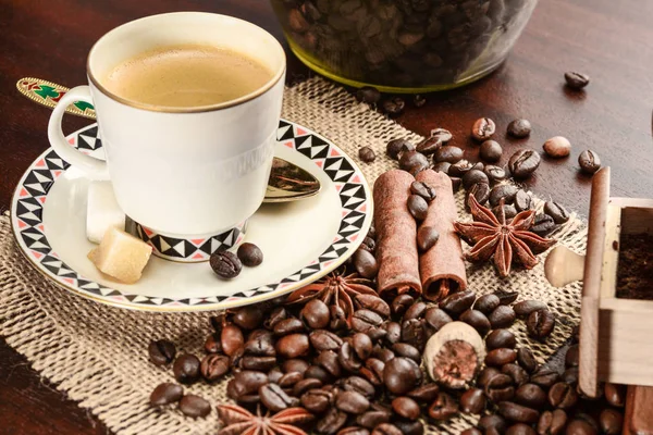 Cup of coffeee with saucer on a jute napkin. Vintage still life on wooden table with sugar, anise, cinnamon, grinder and coffee jar, close up — Stock Photo, Image