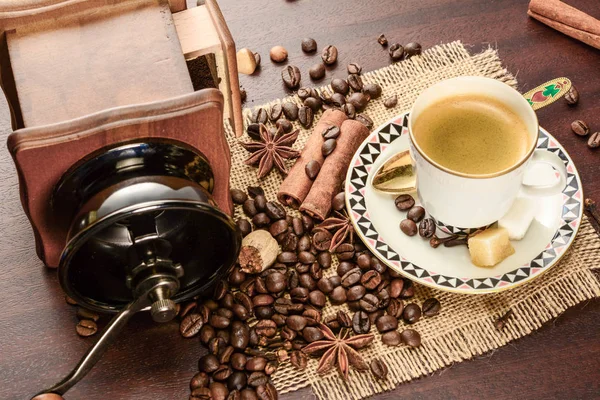 Coffee cup with saucer on a jute napkin. Vintage still life on wooden table with sugar, anise, cinnamon, grinder and dropped coffee beans