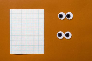 Pair of google eyes are looking at sheet of notebook with copy space for your text, on a brown paper background. Mad funny toys eyes. clipart
