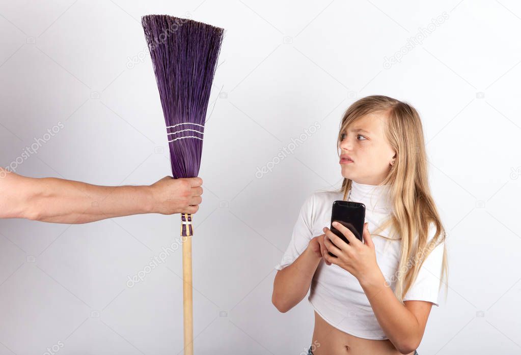 Young girl playing with a smartphone not too impressed by a hand holding a corn broom. Children and housework concept. The smartphone addicting blocking teens from helping in the chore.