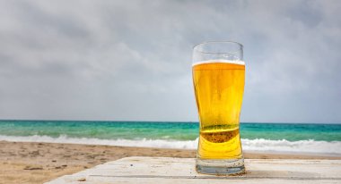 Bardak soluk altın Pilsner Bira bir kumsalı ve mavi yeşil okyanus üzerinde gri bulutlu bir günde bakan bir rustik ahşap masa üzerinde deniz kenarında