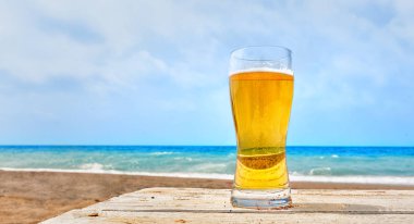 Cam bir tropikal kumlu plaj ve sakin mavi okyanus yaz tatillerini kavramsal bakan bir rustik ahşap masa üzerinde deniz duran, soluk altın Pilsner Lager