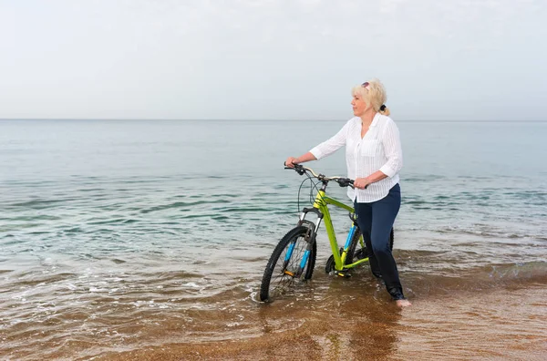 Blond woman wading through the sea pushing a bicycle through the surf as she looks out over the calm ocean
