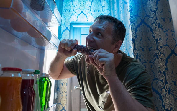 饥饿的男人吃零食从他的冰箱咬到整个香肠看着从敞开的门内寻找 — 图库照片