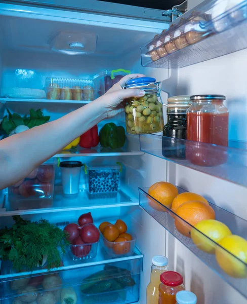 Person reaching for jar with green olives in open fridge full of food