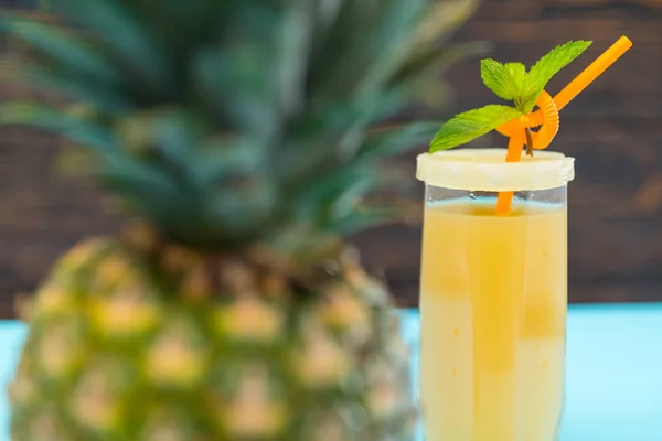 View past a pineapple of a glass of freshly squeezed juice or smoothie served with a straw garnished with a slice of canned pineapple and mint