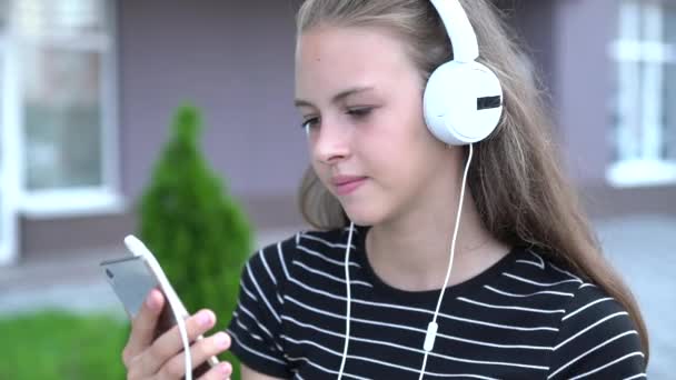 年轻的妇女与淡棕色头发和白色耳机笑着 而看着她的智能手机 — 图库视频影像