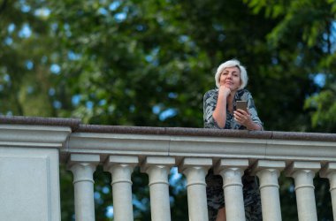 Gülümseyen kadın yapraklı yeşil ağaçlar çerçevede bir kameraya bakıyor bir parapetin üzerinden eğilerek