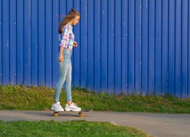 İnce trendy genç kadın akşam ışık renkli mavi duvar önünde bir yol boyunca kasabada kaykay