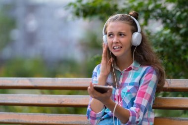 Genç kadın o cep telefonunu açık havada yeşil karşı ahşap Bank müzik dinler gibi dehşete bir yüz çekerek