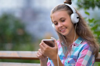 Gülümseyen genç kadın açık havada stereo kulaklıklar müzik dinlemek oturur gibi cep telefonu yeni bir müziği seçme