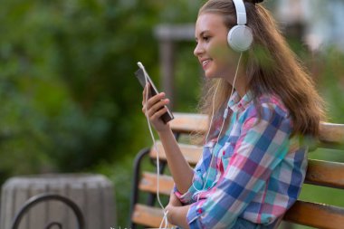 Oturan gülümseyen genç kız açık havada stereo kulaklık cep telefonu kullanarak ahşap bir bankta onu müzik keyfi
