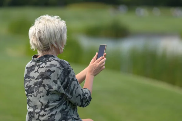 老妇人站在户外与她的手机在她的手中阅读一条短信对绿色草坪背景 — 图库照片