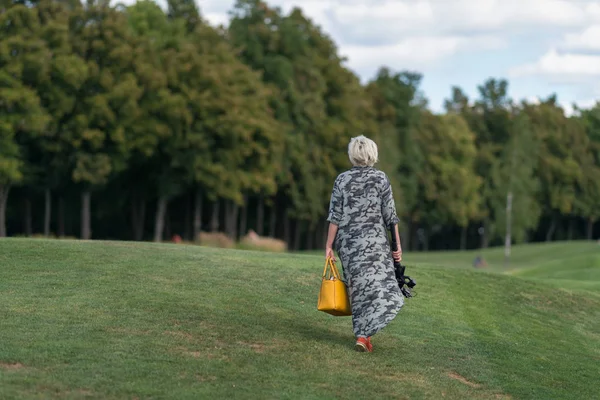 きちんとカット草上林地における背面に向かって堤防の上を歩いて三脚にハンドバッグとカメラを運ぶ長いドレスを着た女性 — ストック写真