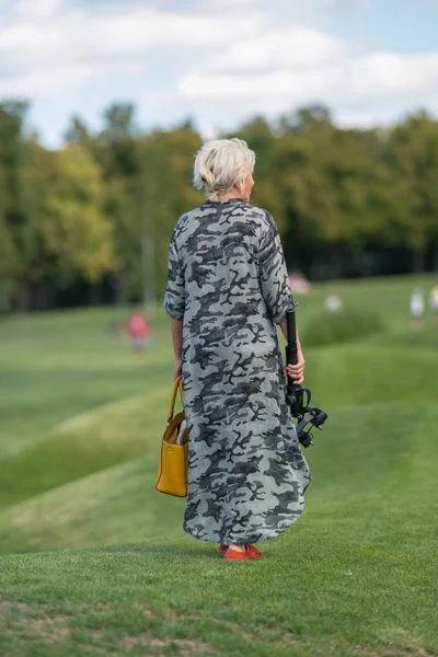 背面に接続されたカメラとハンドバッグ三脚を運ぶ芝生の上を歩く女性 — ストック写真