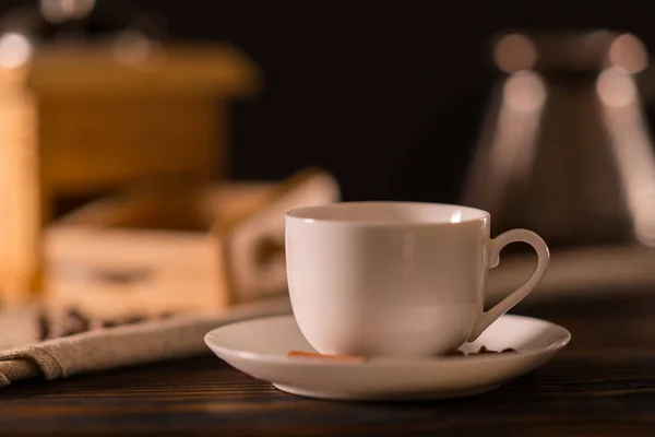 後ろにぼやけているコーヒーミルの低角度側ビューを間近に木製のキッチン テーブルの上のプレーン白いコーヒー カップ — ストック写真