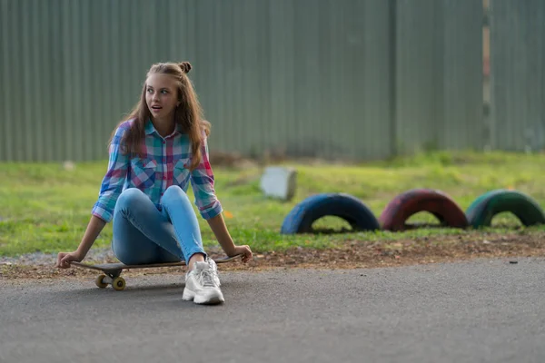 Young woman sitting waiting on a skateboard on an urban road looking to the side expectantly watching for someone to arrive with copy space