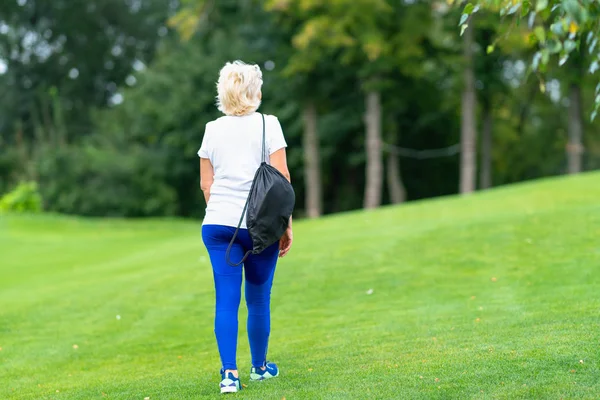 遠くにある木々 に向かって公園の緑の草を離れて歩いて彼女の肩越しにスポーツ バッグを運ぶフィットの女性 — ストック写真
