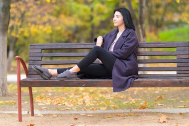 Genç kadın bir rustik ahşap Bank düşünceli kapalı arkasında renkli yeşillik ile sonbaharda tarafına bakarak bir parkta üzerinde rahatlatıcı trendy