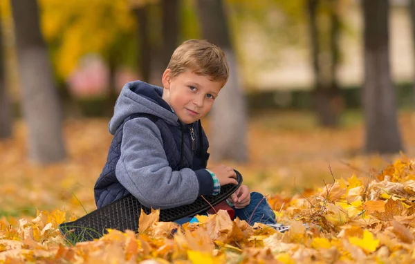 茶色の紅葉公園の中に座っている彼のスケート ボードの笑みを浮かべて少年 — ストック写真