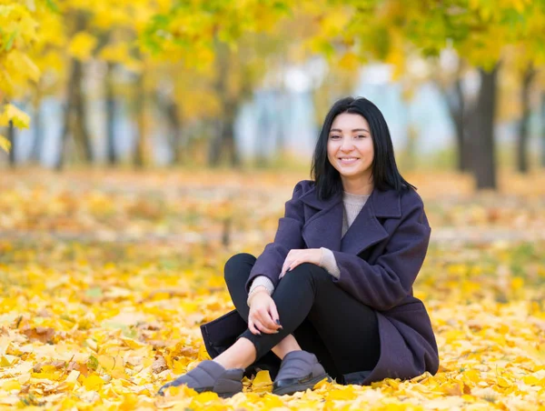 秋の中で座っている魅力的なトレンディな若い女性のカメラで幸せそうに笑って公園の葉します — ストック写真