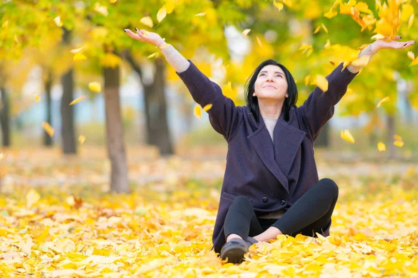 当她坐在公园里的地上时 无忧无虑的年轻女子在庆祝秋天的季节 在空中抛着五颜六色的黄叶 — 图库照片