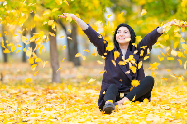Happy young woman throwing yellow autumn leaves into the air as she sits on the ground in a park
