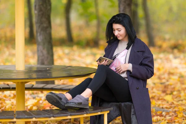 若い女性の笑顔で携帯電話を使用してカラフルな紅葉と公園内の円形テーブルでリラックス — ストック写真