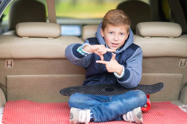 魅力的な小さな男の子のフレームを作る車のトランクに座ってカメラに微笑むように彼の指でジェスチャー — ストック写真