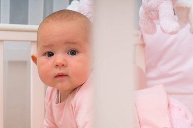 Şirin bebek kız beşik, kamera, pembe tişörtlü beyaz parmaklıklar arkasında oturan bakarak. Yakın çekim portre kopya alanı ile
