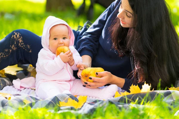 カラフルな秋の公園で草で敷物の上彼女の母と彼女が座っているように熟した黄金のリンゴをかむ女の子赤ちゃん葉します — ストック写真