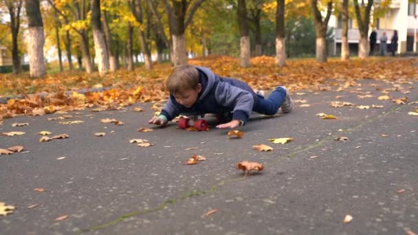 小男孩试图骑滑板 躺在肚子上 用手推离地面 从公园人行道上的地面上看 有黄色的秋叶 — 图库视频影像