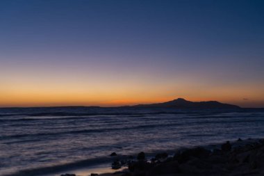 Bir tropikal okyanus ve kayalık bir sahilden bir sakin mavi okyanus üzerinde görüntülenen uzak dağ üzerinde renkli turuncu günbatımı