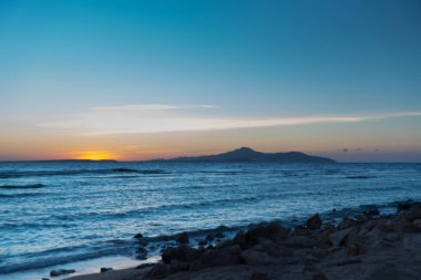Okyanusu günbatımı turuncu gökyüzü ve açık mavi gökyüzü altında ön planda kayalık sahil şeridi ile
