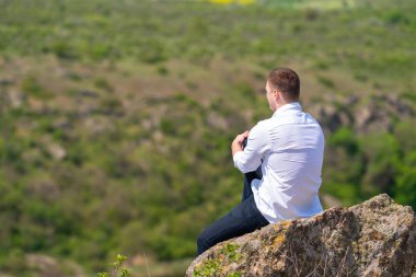 Genç adam yüksek bir kayanın üzerinde oturuyor.
