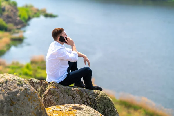 Genç adam bir kayanın üzerinde oturuyor ve cep telefonuyla sohbet ediyor. — Stok fotoğraf