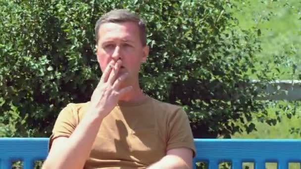 Человек, сидящий на скамейке в парке и курящий — стоковое видео