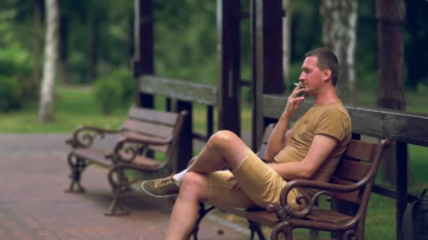 Man seated on a park bench smoking a cigarette — Stock Video