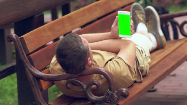 Мужчина отдыхает на скамейке в парке под солнцем — стоковое видео