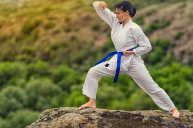 Genç bir kadın dövüş sanatları ya da açık havada bir kayanın üzerinde kickboks yapıyor ve yeşilliğe karşı yan gözle poz veriyor.